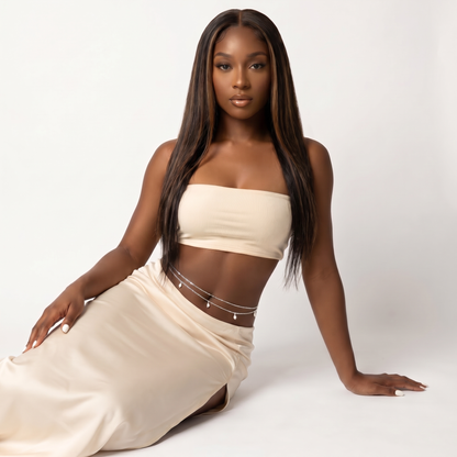 A woman with long, straight dark brown hair models a matching cream-colored two-piece set consisting of a strapless crop top and a high-waisted wrap skirt, accessorized with delicate silver chain details. She is posed on a seamless light gray studio background.
