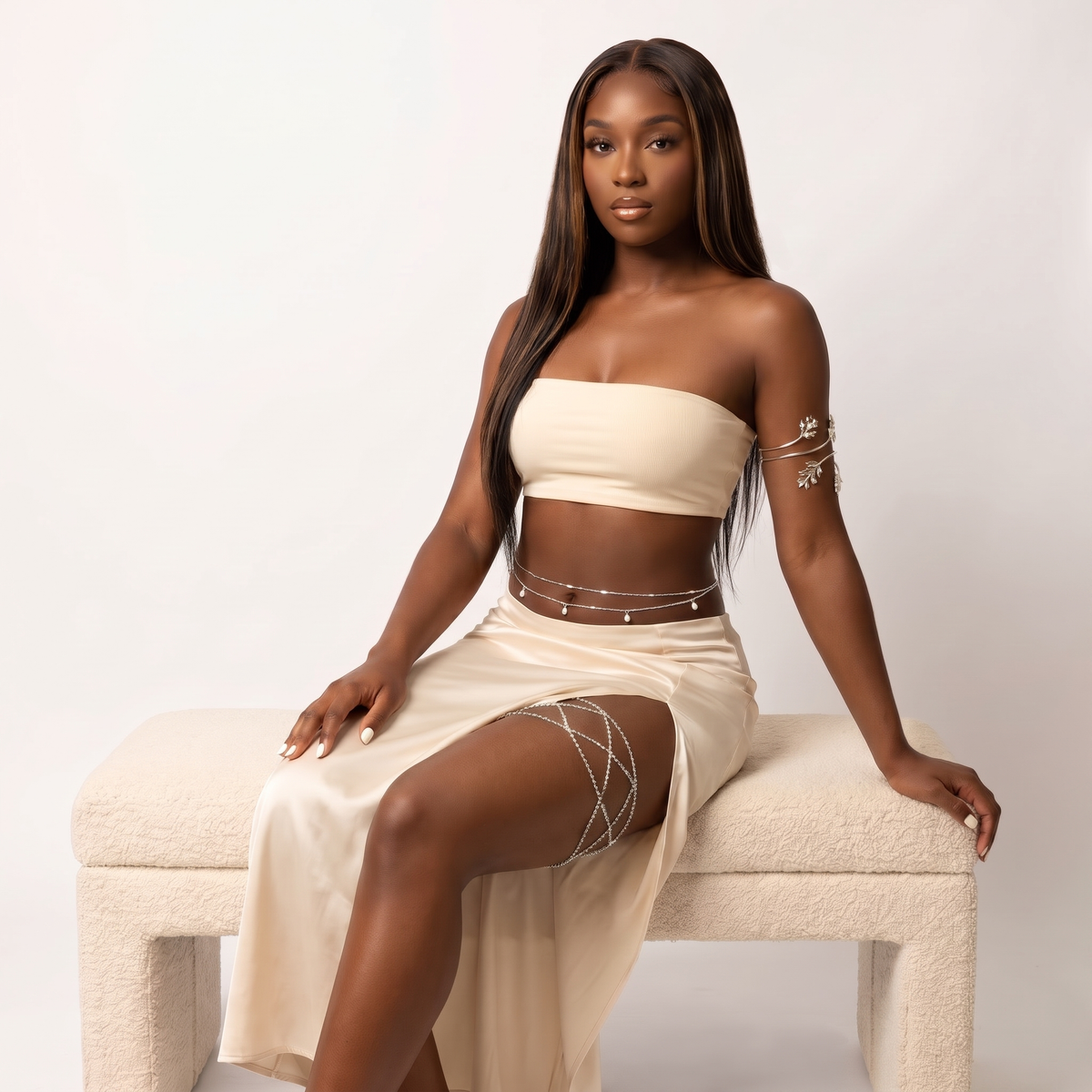 A woman with long dark hair sits on a cream boucle bench wearing a matching cream two-piece set with a strapless bandeau top and a high-slit skirt, accented with silver chain details on her waist, hips, and upper thigh.