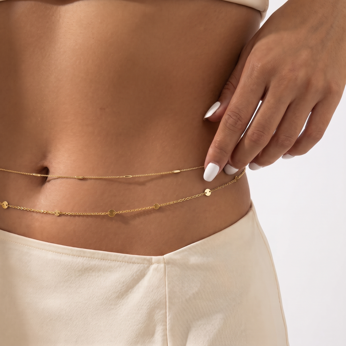 A close-up of a model's midsection wearing a delicate gold double-layered belly chain with small disc charms, paired with cream-colored bottoms.