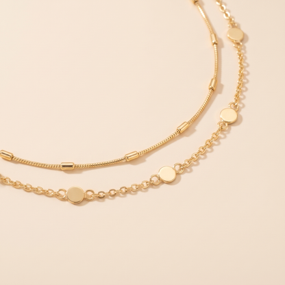 A delicate gold chain necklace with flat circular disc charms, displayed on a warm beige surface with a soft, warm-toned shadow.
