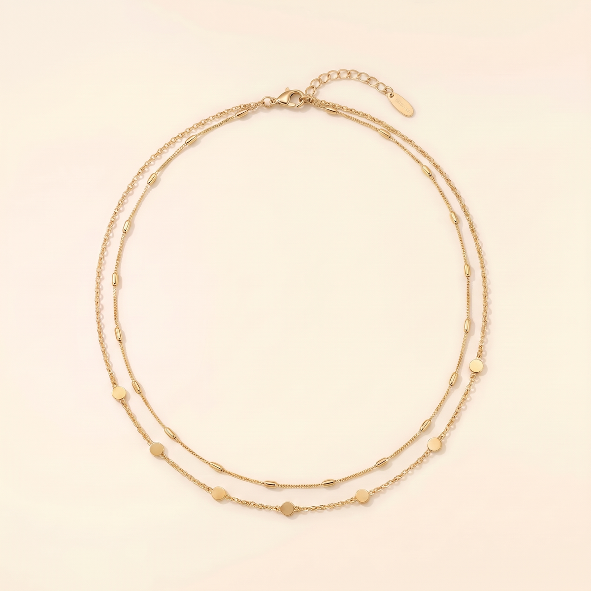 A delicate double-strand gold chain necklace with small, evenly spaced heart-shaped and oval beads, displayed on a warm cream background.