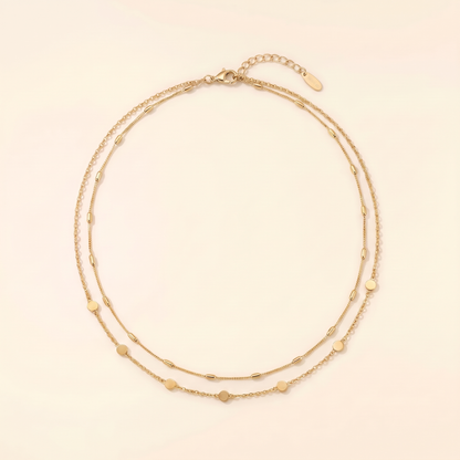 A delicate double-strand gold chain necklace with small, evenly spaced heart-shaped and oval beads, displayed on a warm cream background.