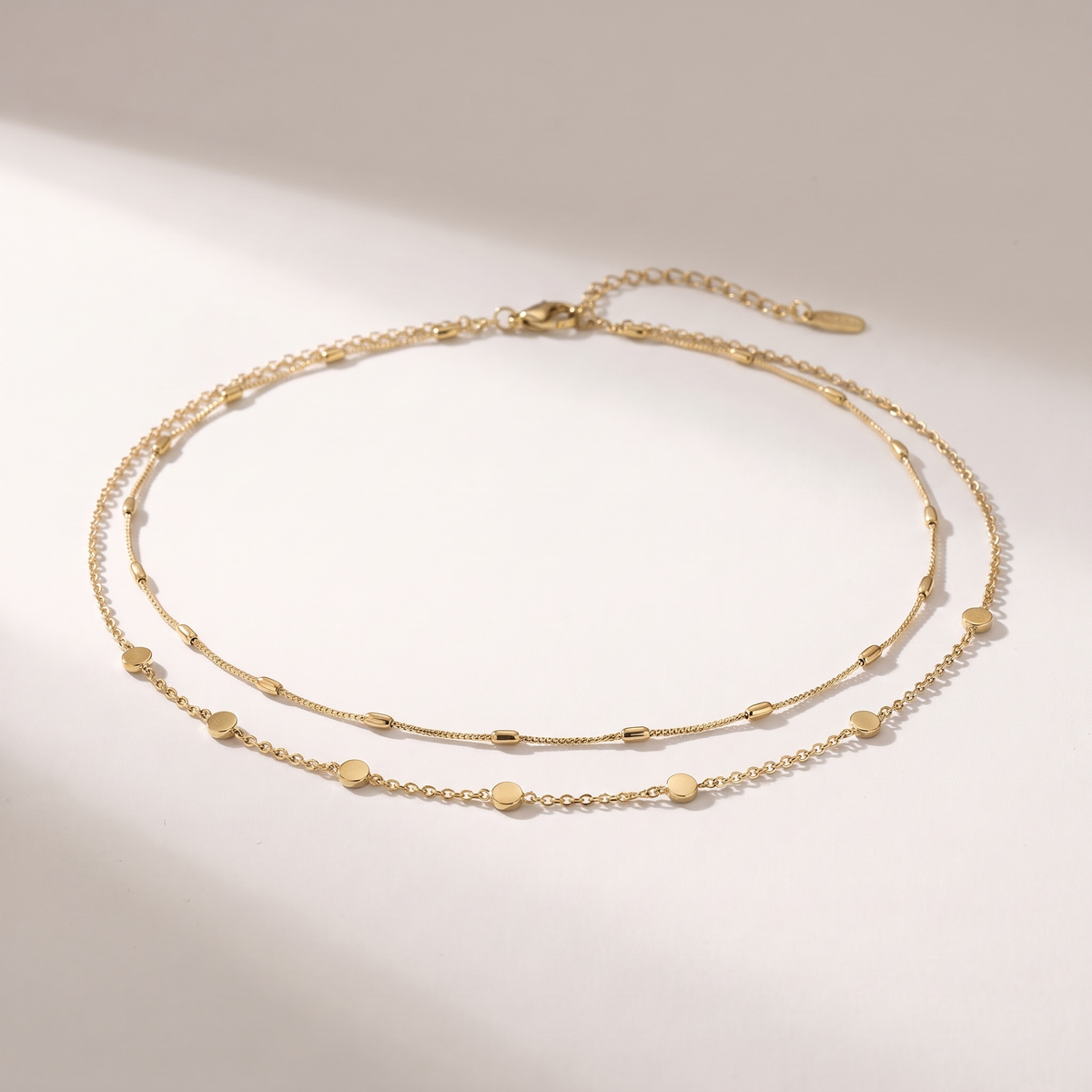 A delicate double-layered gold chain anklet with small flat disc charms, displayed on a clean white surface with soft shadows.